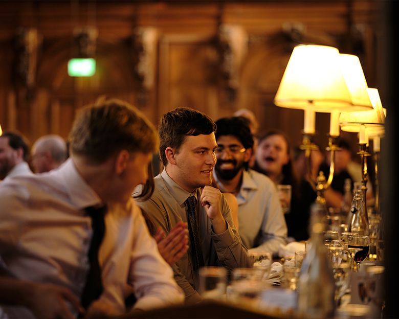 A student sitting at an upscale dinner table listens to a speaker off camera.