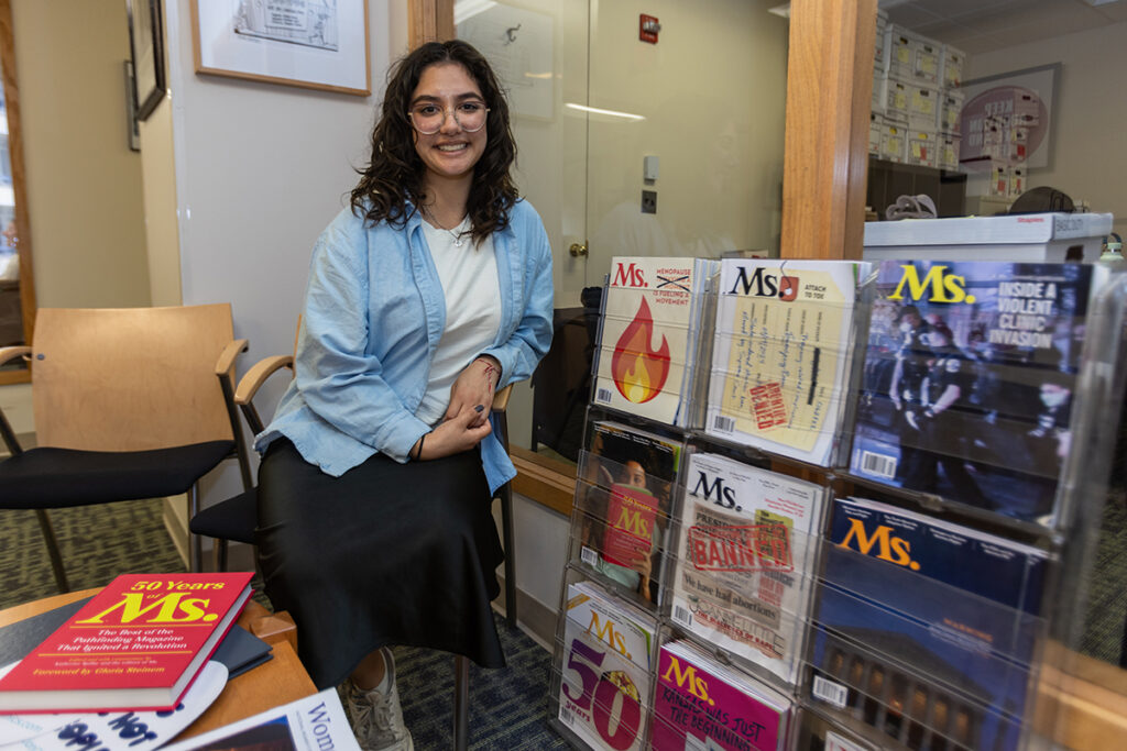 Abby Ramirez sits next to a rack of magazines in an office setting.