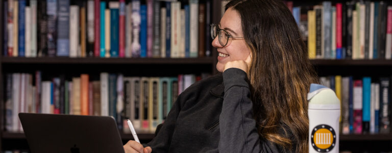 A college student sits at a table and smiles at someone off camera. There is an open laptop and a water bottle on the desk. There is a bookshelf in the background.