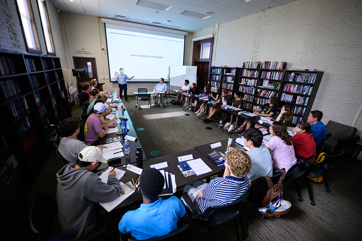 A group of students sit around tables set up in a "U" shape and listen to a speaker who stands in front of a projector.