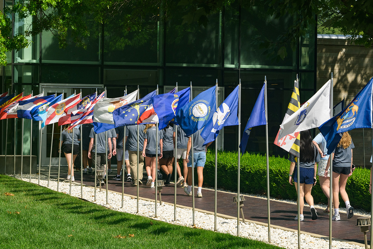 Students walk in a line behind a row of state flags on poles.