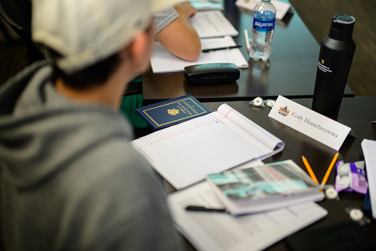 A student sits at a table and listens to an off camera speaker. Focus is on their belongings on the table, including a notebook, the United States constitution, a water bottle, a name tag, and a few pencils.