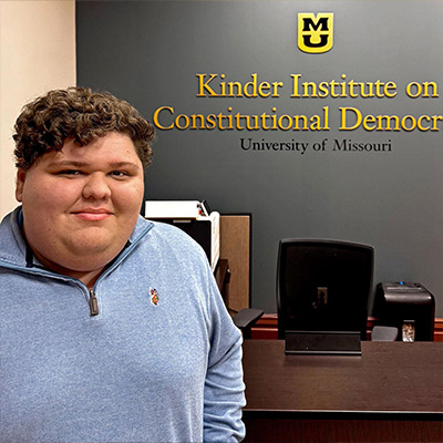 A student stands in front of a desk and a wall with the Kinder Institute logo.