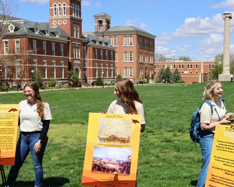 Three college students present posters about the Santa Fe Trail outdoors on campus.
