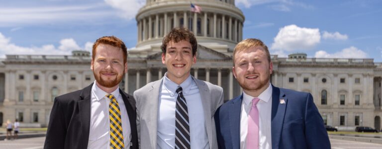 Three young men in suits stand in front of the US Capitol.