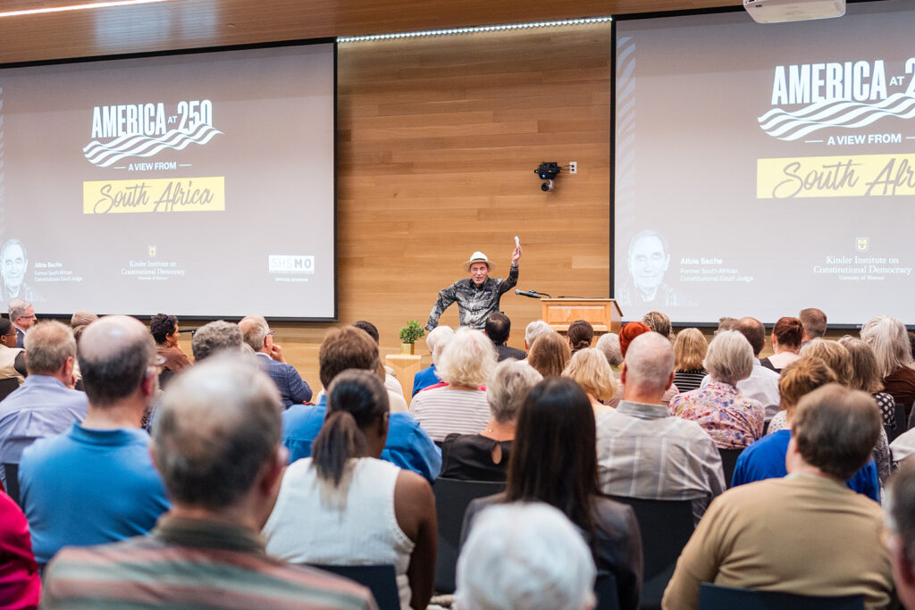 A speaker talks to an audience. Projectors on the wall show "America at 250: A View from South Africa."
