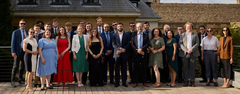 A group of nicely dressed grad students stand and smile together on a patio at Oxford.