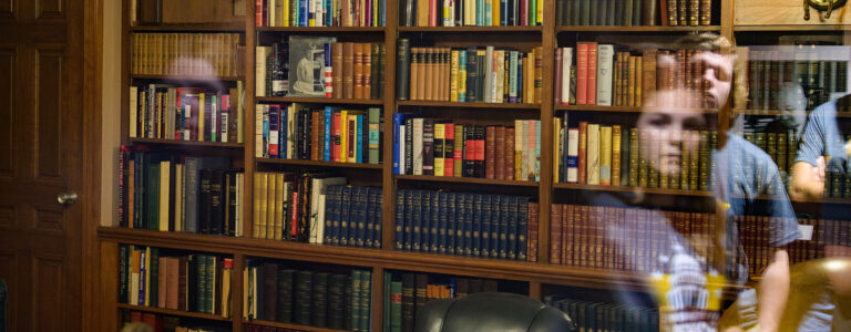 Library bookshelves seen through glass. Reflections of students are visible on the right side of the image.