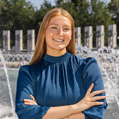 Kate Karpinski smiles for the camera in front of a fountain with her arms crossed.