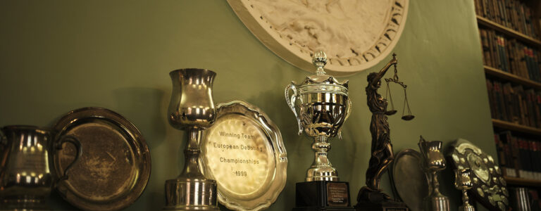 Trophies sit on a shelf in front of a green wall. A plate in the center says 