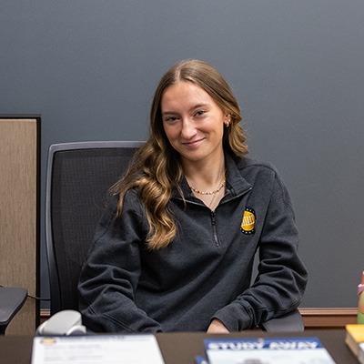 A student sits behind a desk and smiles.