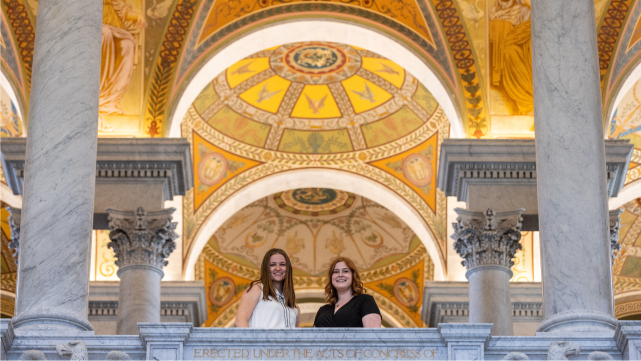 Two students smile for the camera. They stand above the camera, behind a railing, with a decorative arched ceiling in the background.