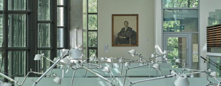 A glass study room with a lattice of desk lamps and a black and white portrait on the wall.