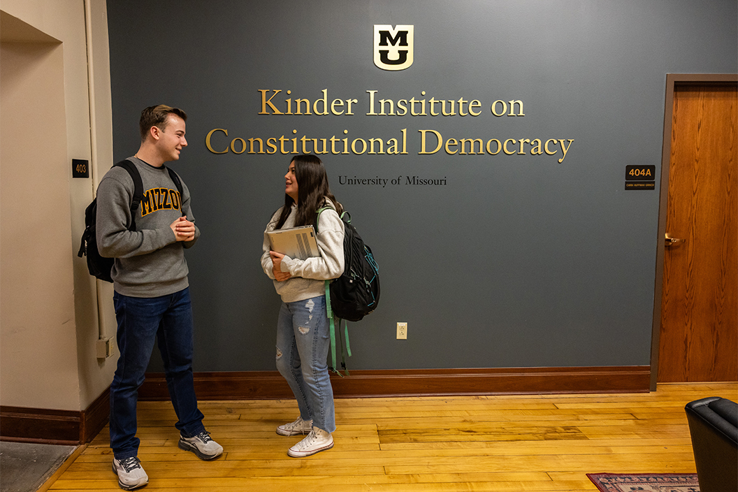 Two college students chat in front of a wall sign with the Kinder Institute logo.