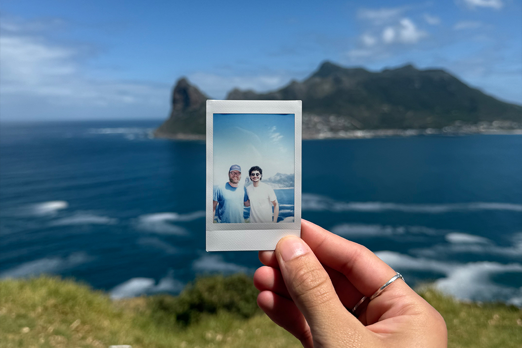 A hand holds up a polaroid photo of two smiling students in front of the ocean.