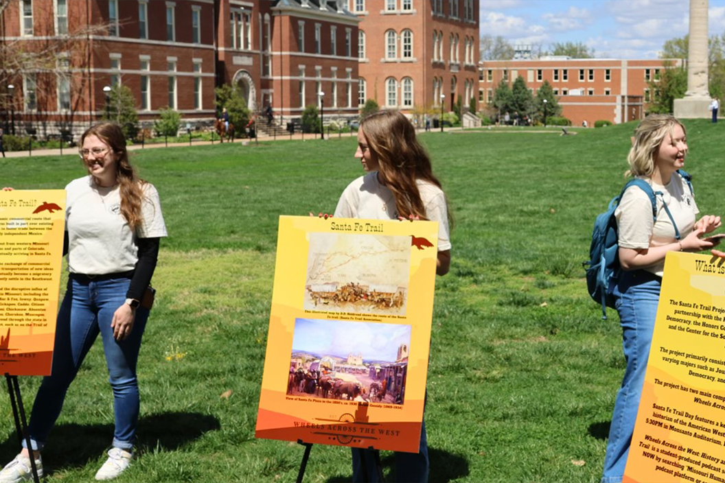 Three college students present posters about the Santa Fe Trail outdoors on campus.