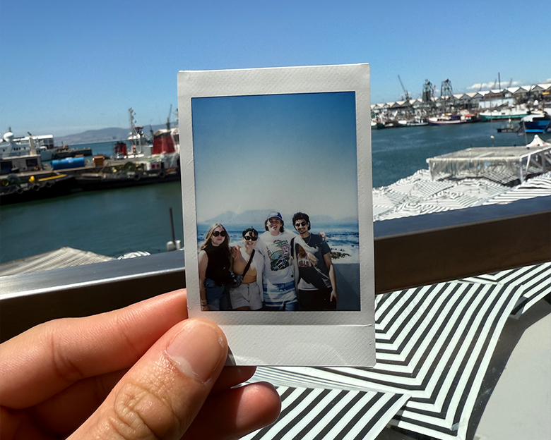 A student holds up a polaroid in front of a marina. The polaroid shows four students smiling for the camera in front of the ocean.