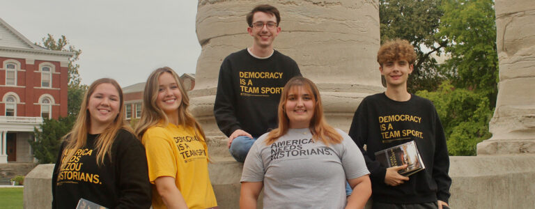 Five college students outside, wearing black and white shirts that say phrases like 