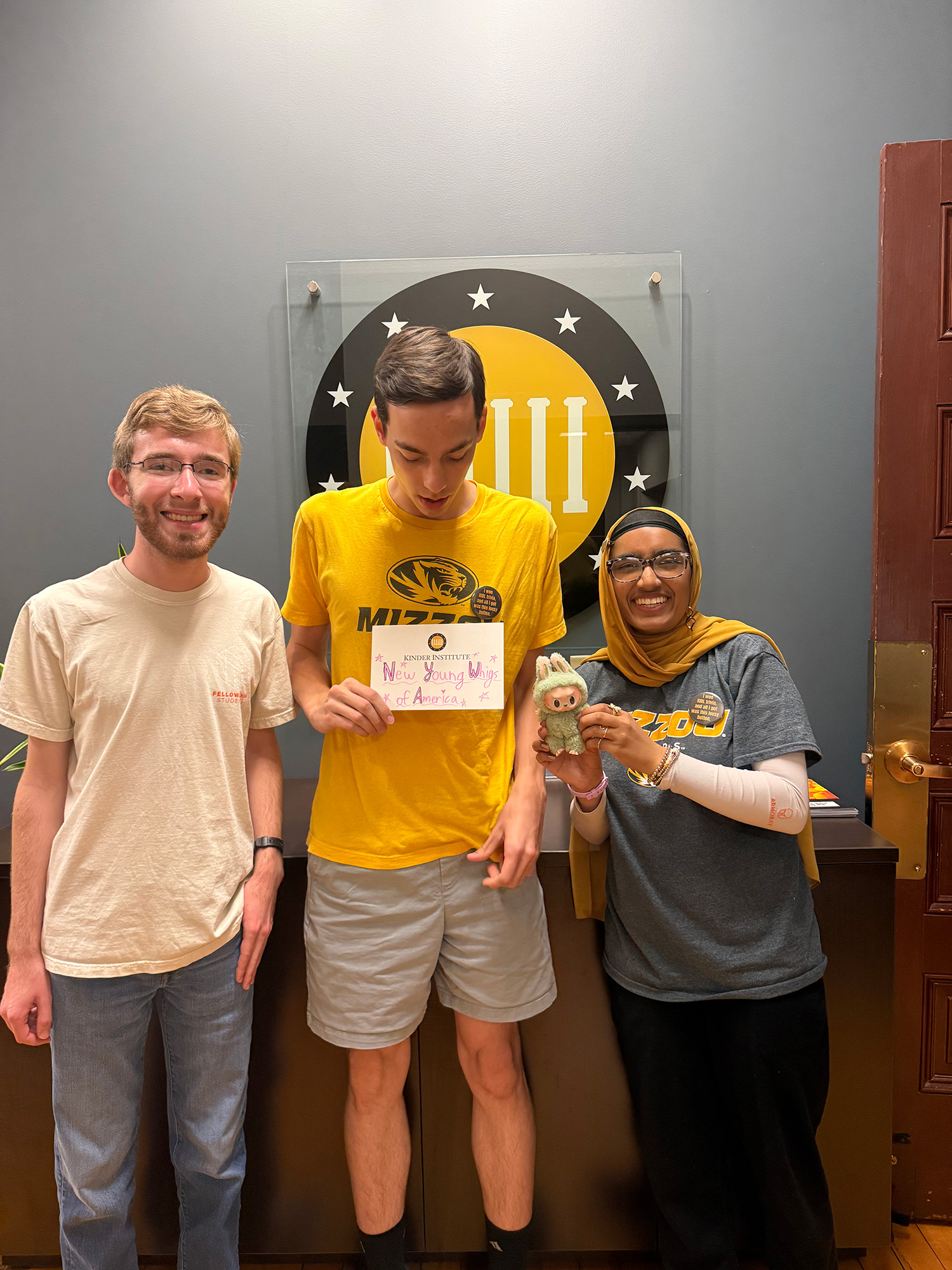 Three students stand and smile for the camera. The student in the middle holds a piece of paper that says "New Young Whigs of America"