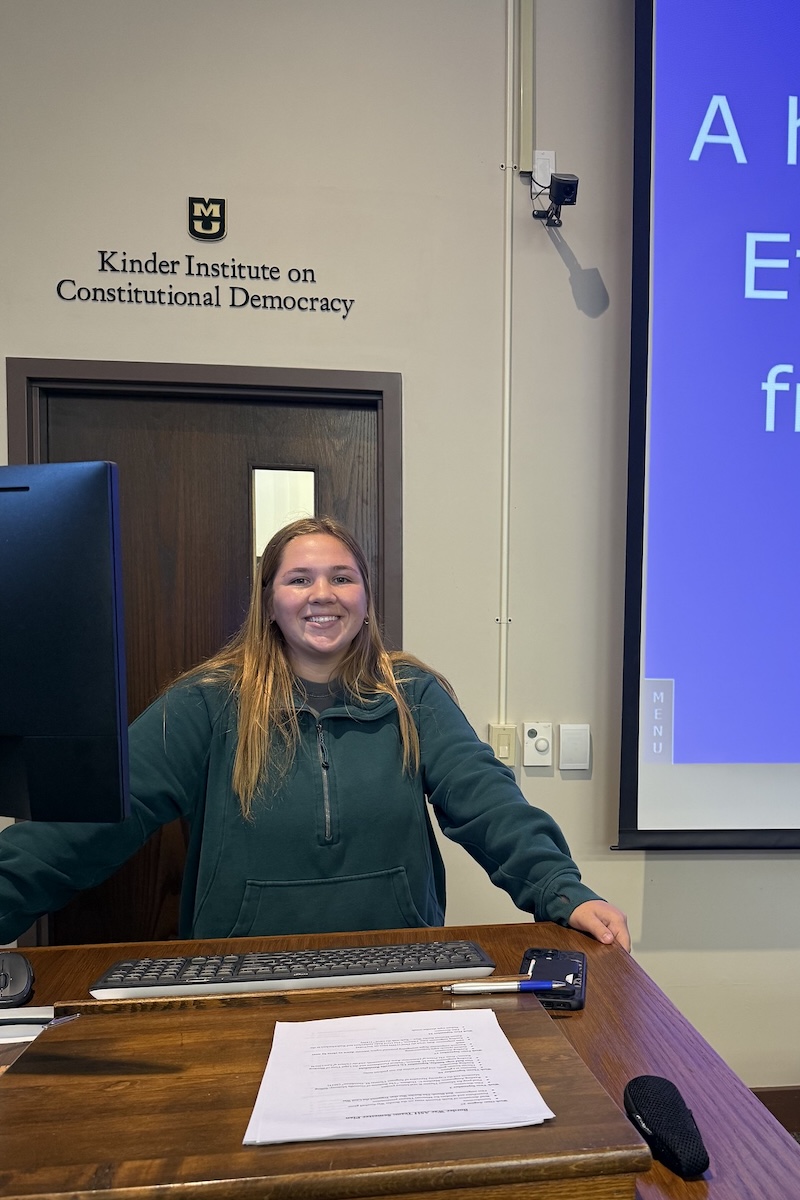 A college student stands at the front of a classroom, managing the projector screen