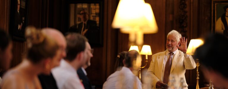 A man in a light suit gives a toast in a historic dining hall