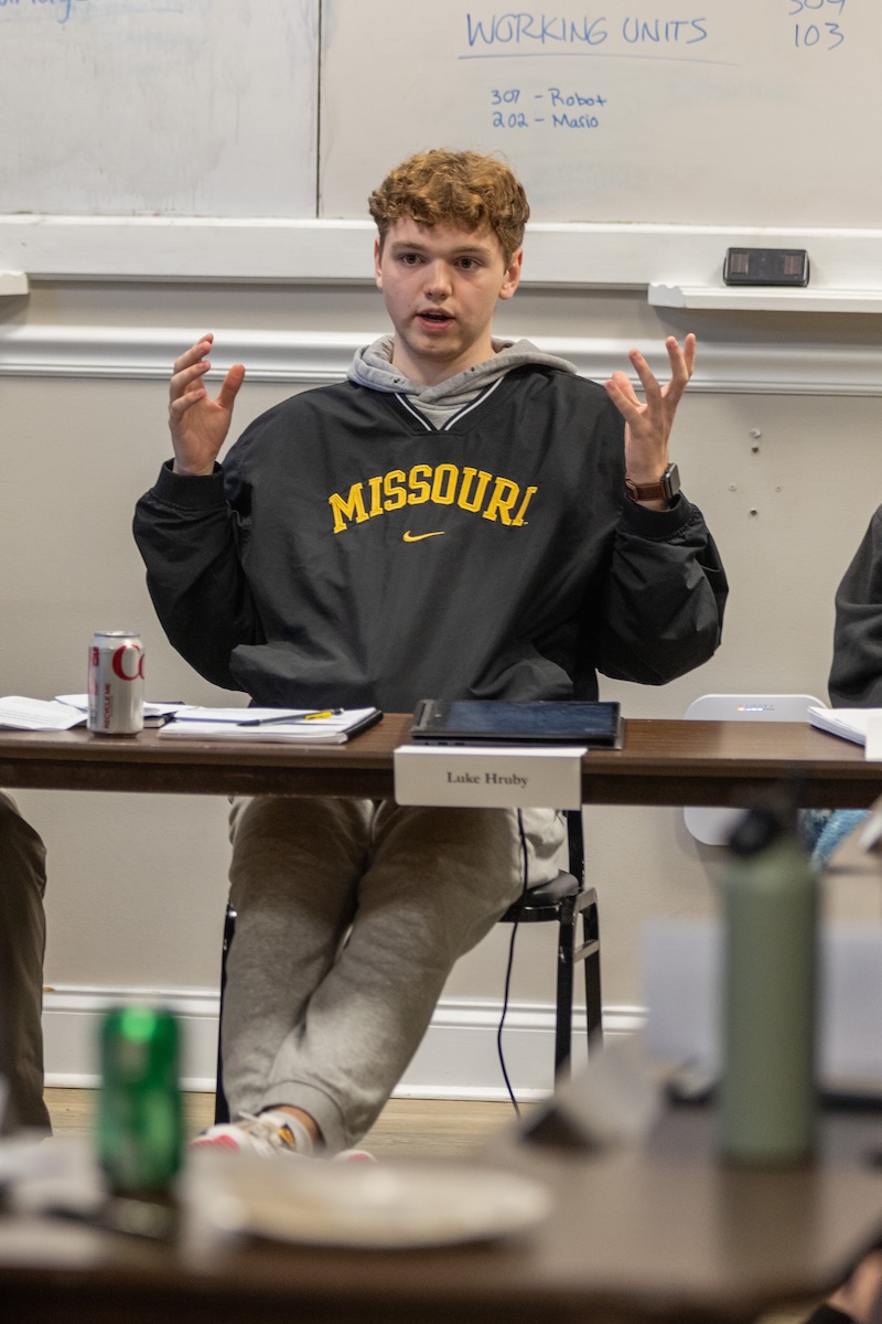 A student in a college classroom gesticulates with his hands while making a point. 