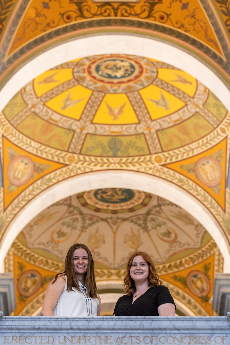 Two students stand on the top balcony of the Library of Congress, the dome's murals illuminated in the background