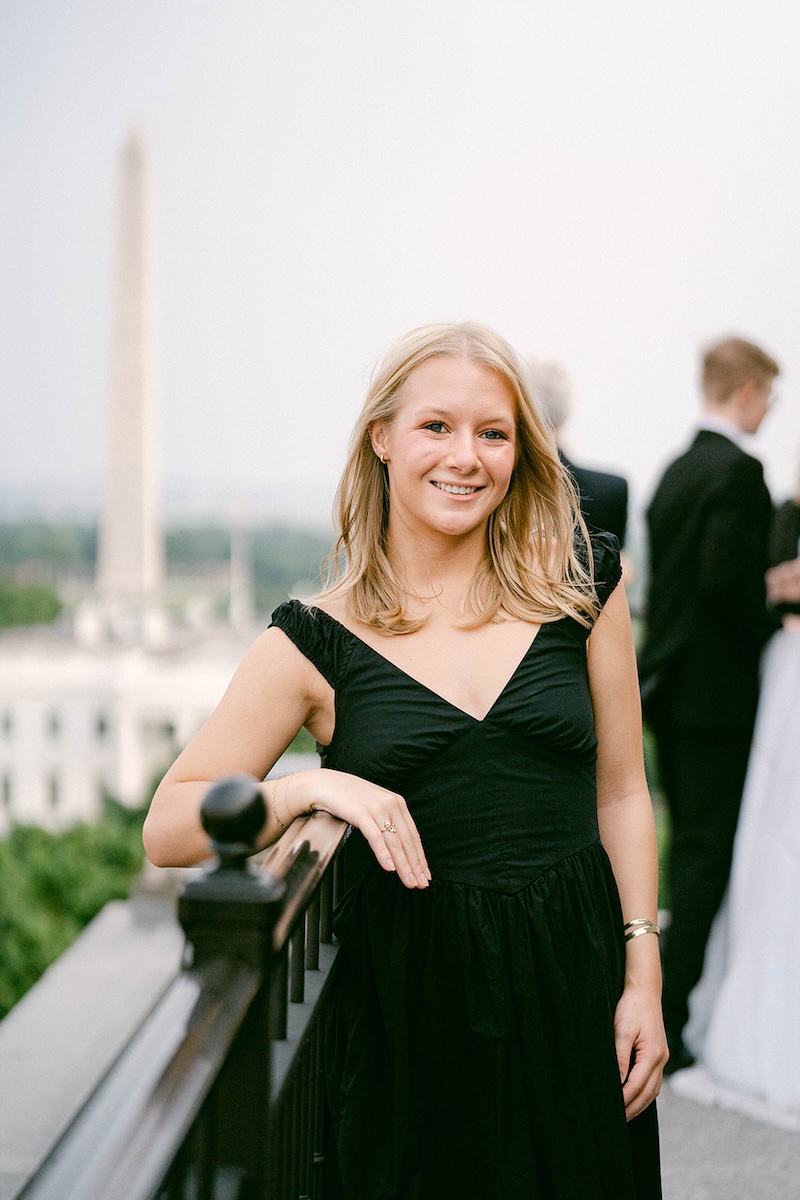 A college student in formal ware stands on the rooftop balcony of the Hay-Adams Hotel, the Washington Monument in the distance behind her. 