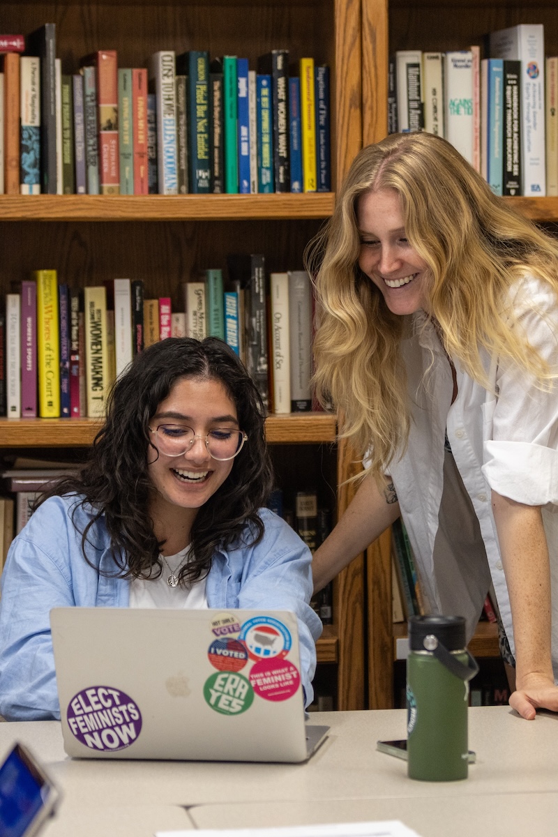 A college intern works at her laptop while her employer looks on happily.
