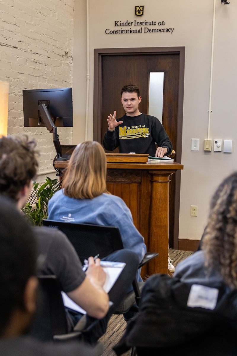 A college student delivers a presentation to his peers from a podium at the front of a classroom