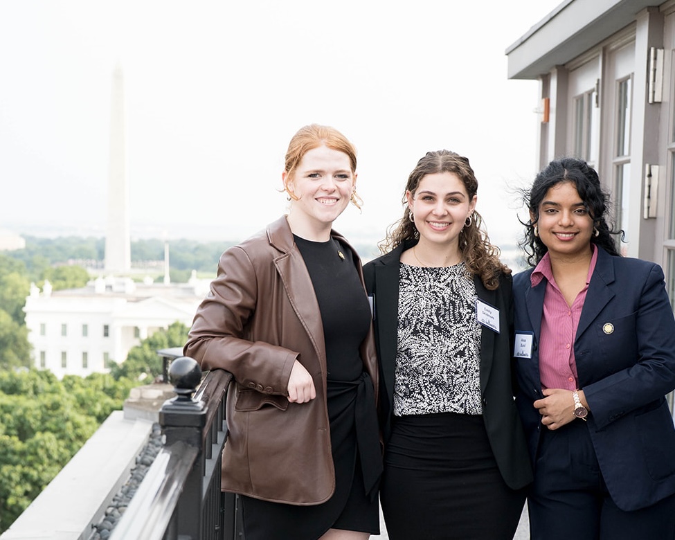Three students stand on a rooftop balcony, the Washington Monument's obelisk in the distance behind them.