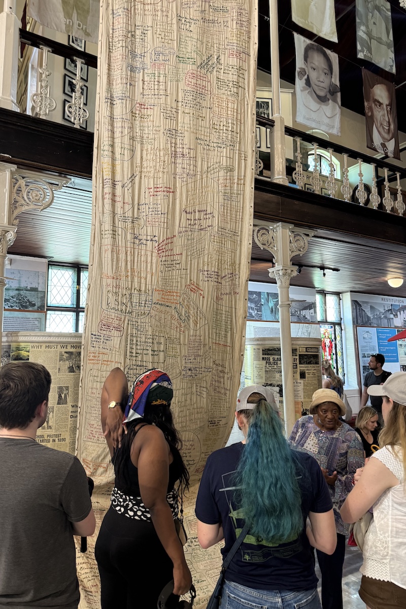 Students admire an illustrated, ceiling to floor tapestry at Cape Town's District Six Museum.