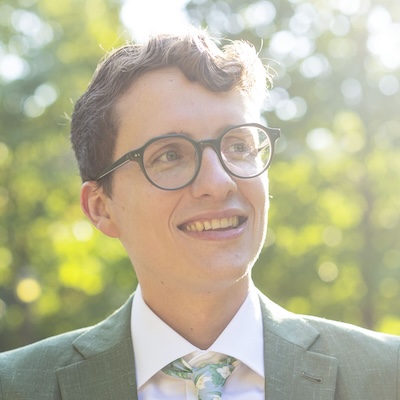 A man with glasses stares wistfully to his left while standing in front of a forest.