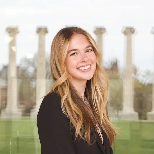 A young woman with blonde hair poses on the veranda of Jesse Hall, a reflection of Mizzou's Columns behind her.