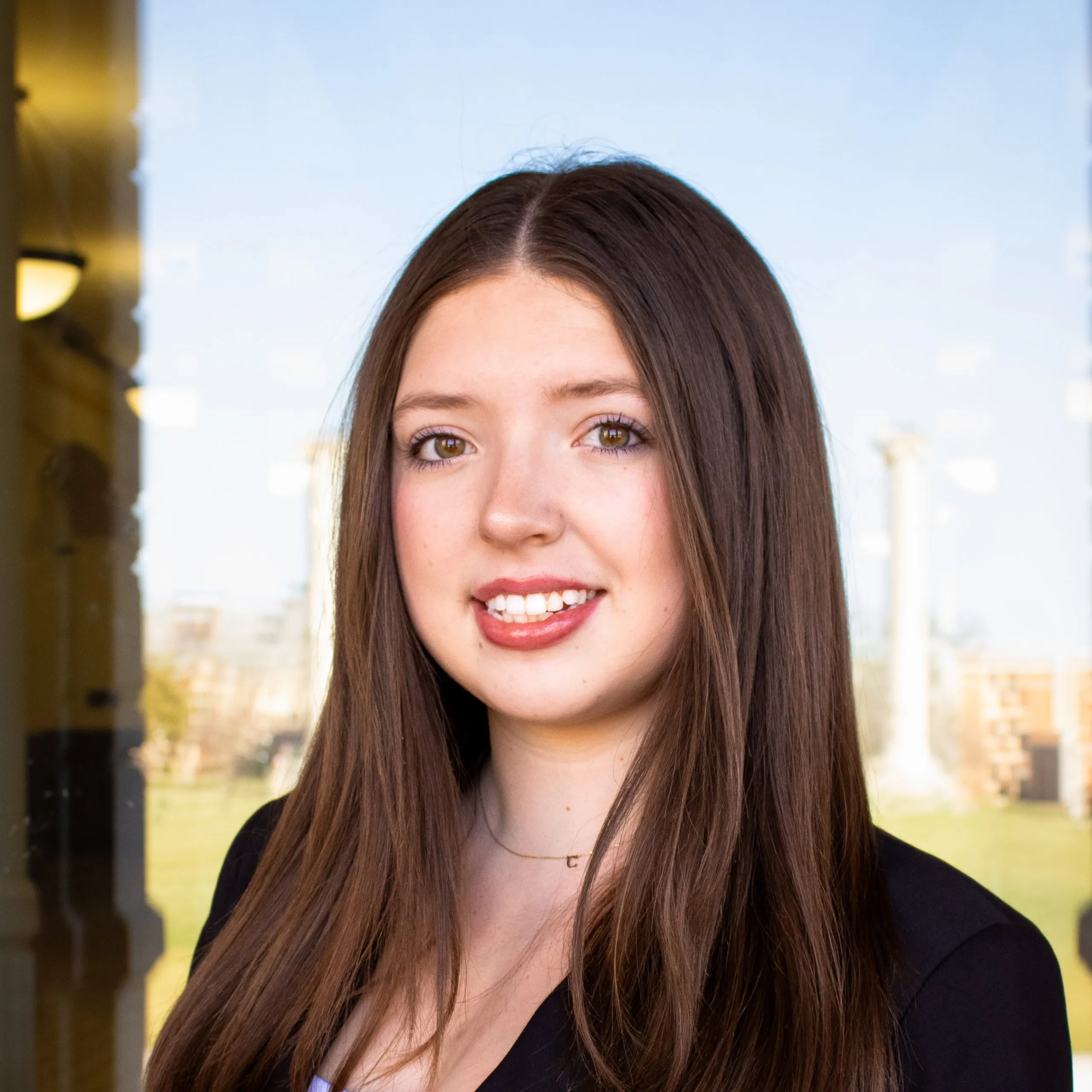 A young woman with brown hair poses for a headshot, the Mizzou Columns reflected in the window behind her