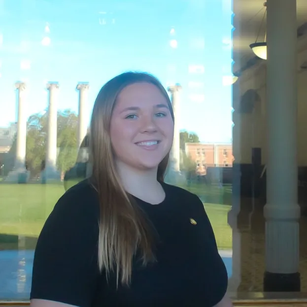 A young woman in a black shirt poses for a headshot, Mizzou's Columns reflected in the window behind her.