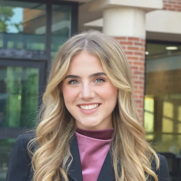 A young woman with blonde hair poses for a headshot in front of a university building.