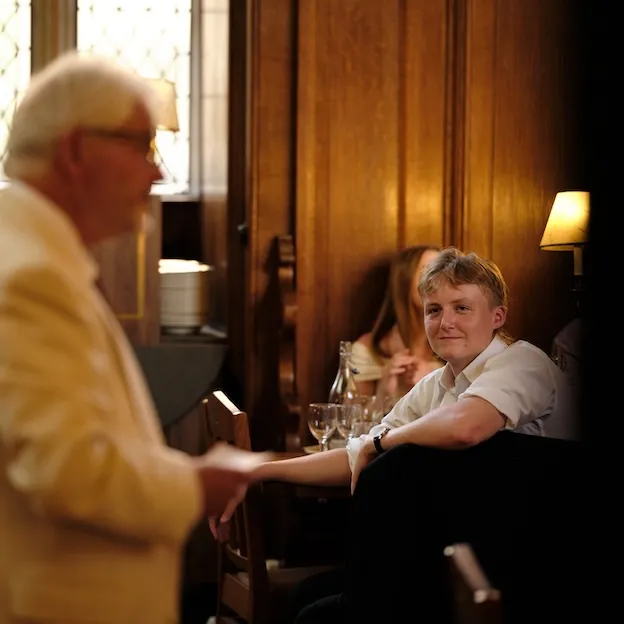 A young man with blonde hair listens on at a dinner lecture in an Oxford dining hall.