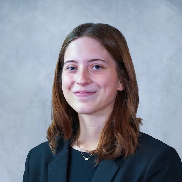 A young woman with a dark blue blazer poses for a headshot in front of a gray backdrop