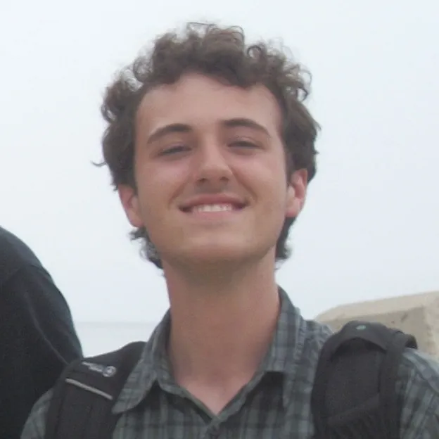 A young man with short brown hair smiles atop a hill