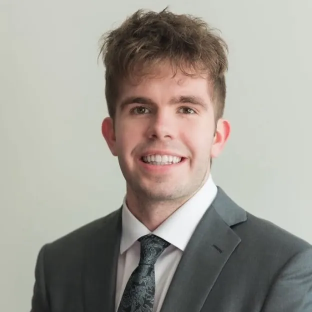 A young man in a gray suit poses for a professional headshot in front of an off-white backdrop