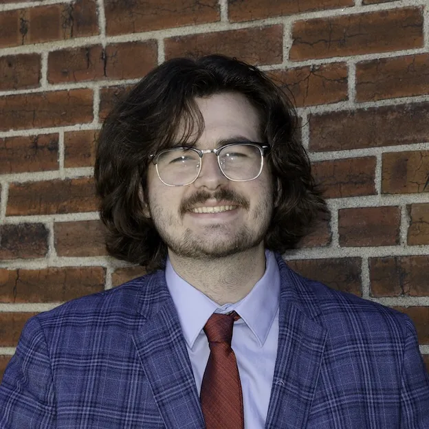 A young man in a blue plaid sportscoat poses for a headshot in front of a brick wall