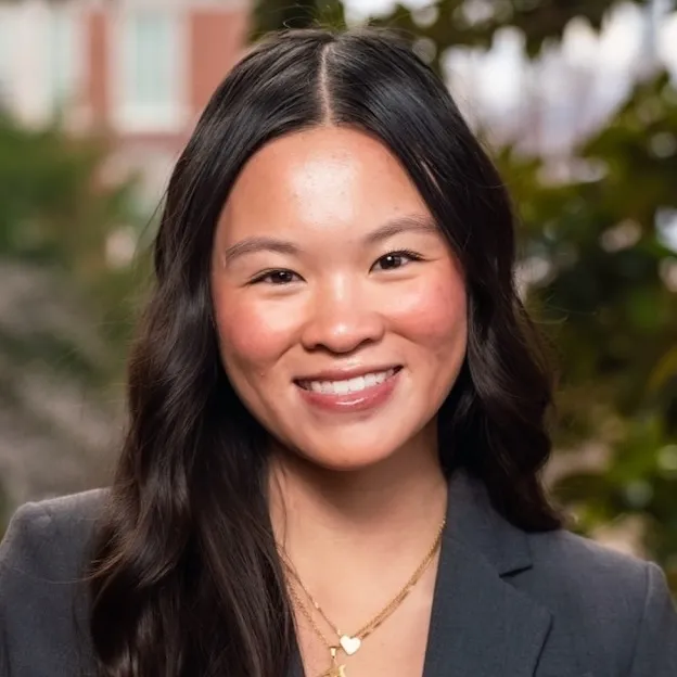A young woman with dark hair poses for a professional headshot, foliage faintly in the background