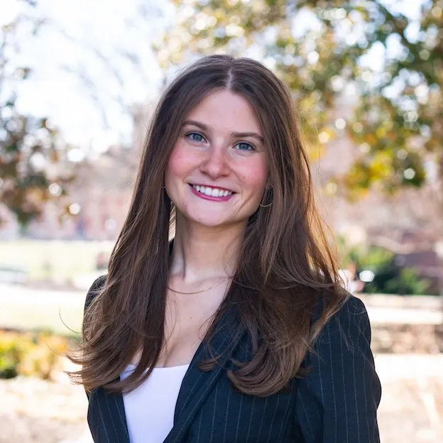 A young woman in a blue jacket poses for a professional headshot, trees blurred in the background.