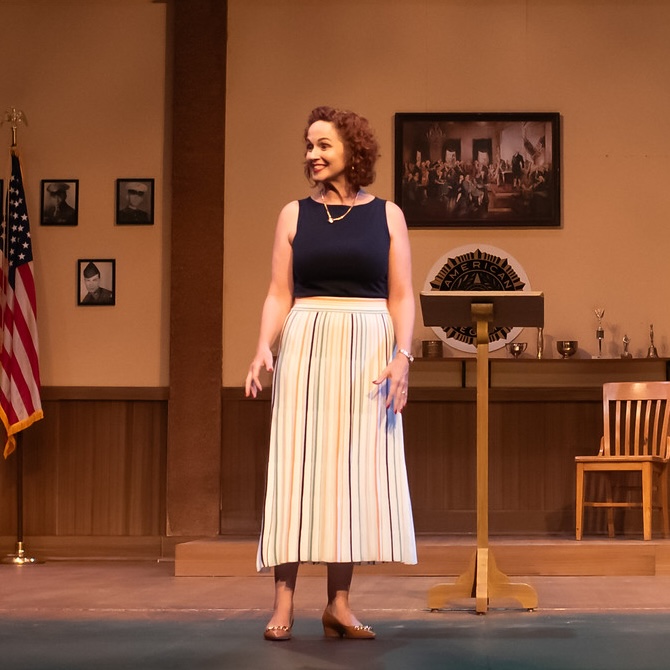 An actress stands at the front of the stage in a performance of What the Constitution Means to Me, globe and American flag in the background.