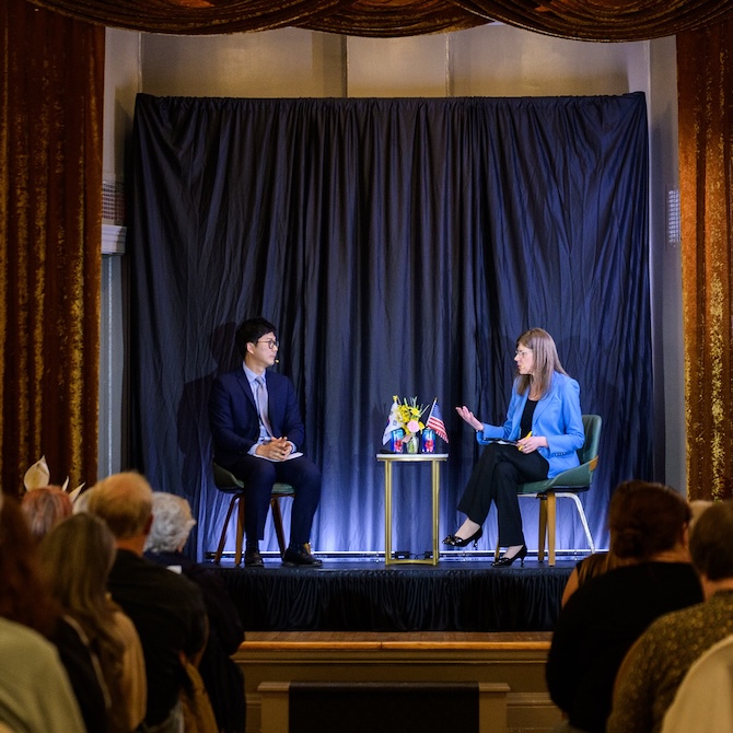 Two scholars converse on stage in a crowded ballroom.