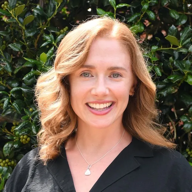A woman with a necklace smiles in front of a backdrop of foliage
