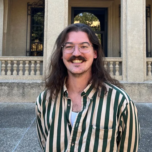 A mustachioed man with a striped shirt stands before the stone edifice of a university library