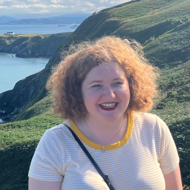 A woman smiles on in rolling hills, a small seaside village in the background