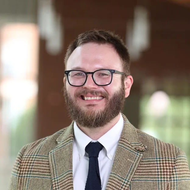 A bearded man in a checked sport coat and glasses smiles for the camera